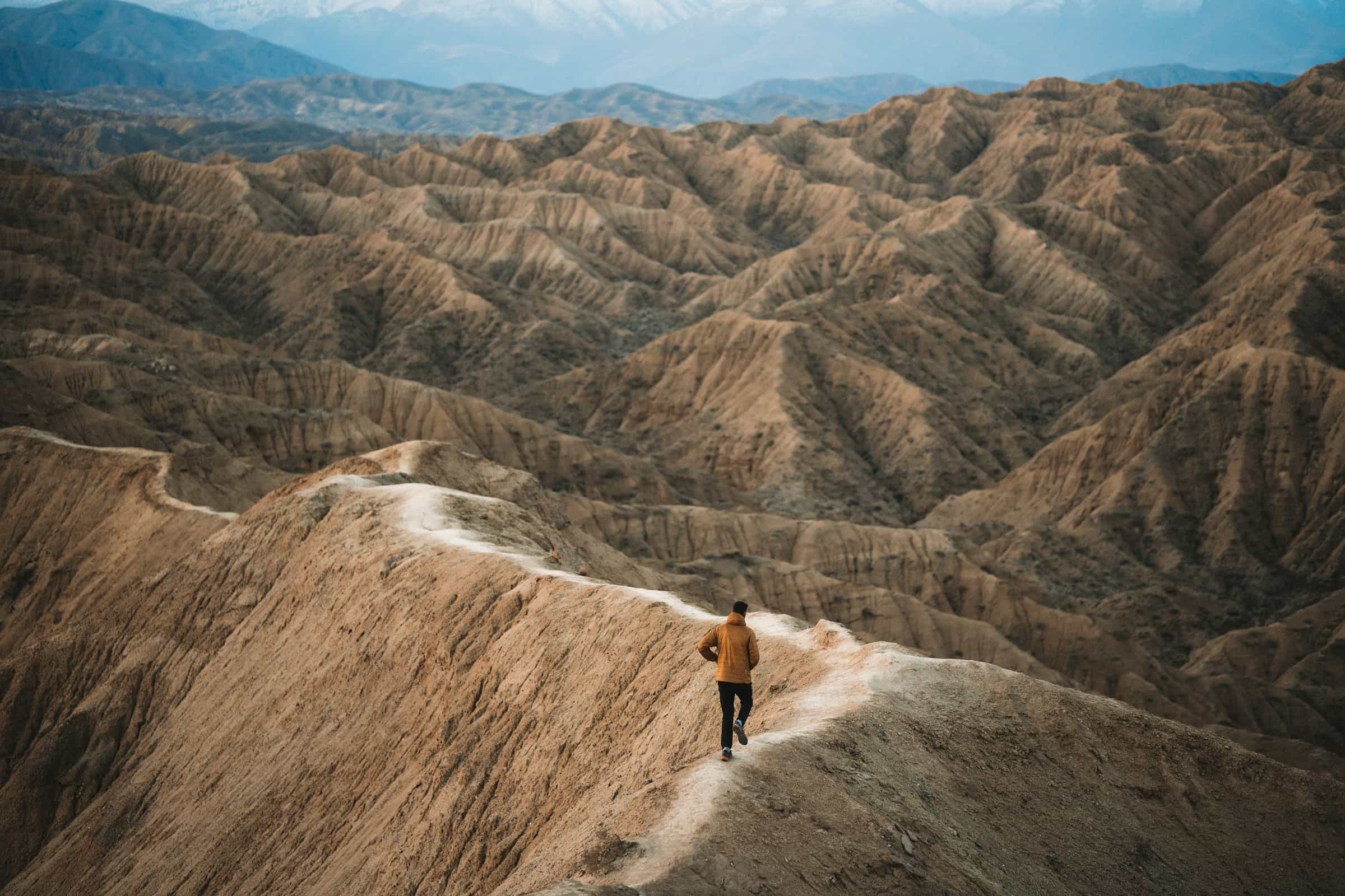 Runner on a narrow trail winding along a ridge through desert badlands